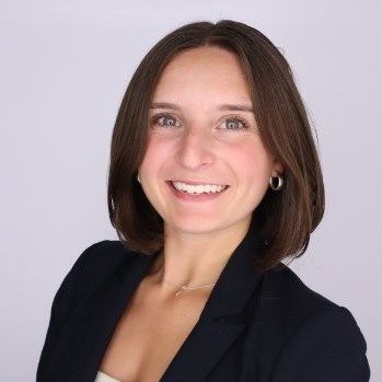 Young woman with shoulder length dark hair, smiling, wearing a dark suit jacket and light top.